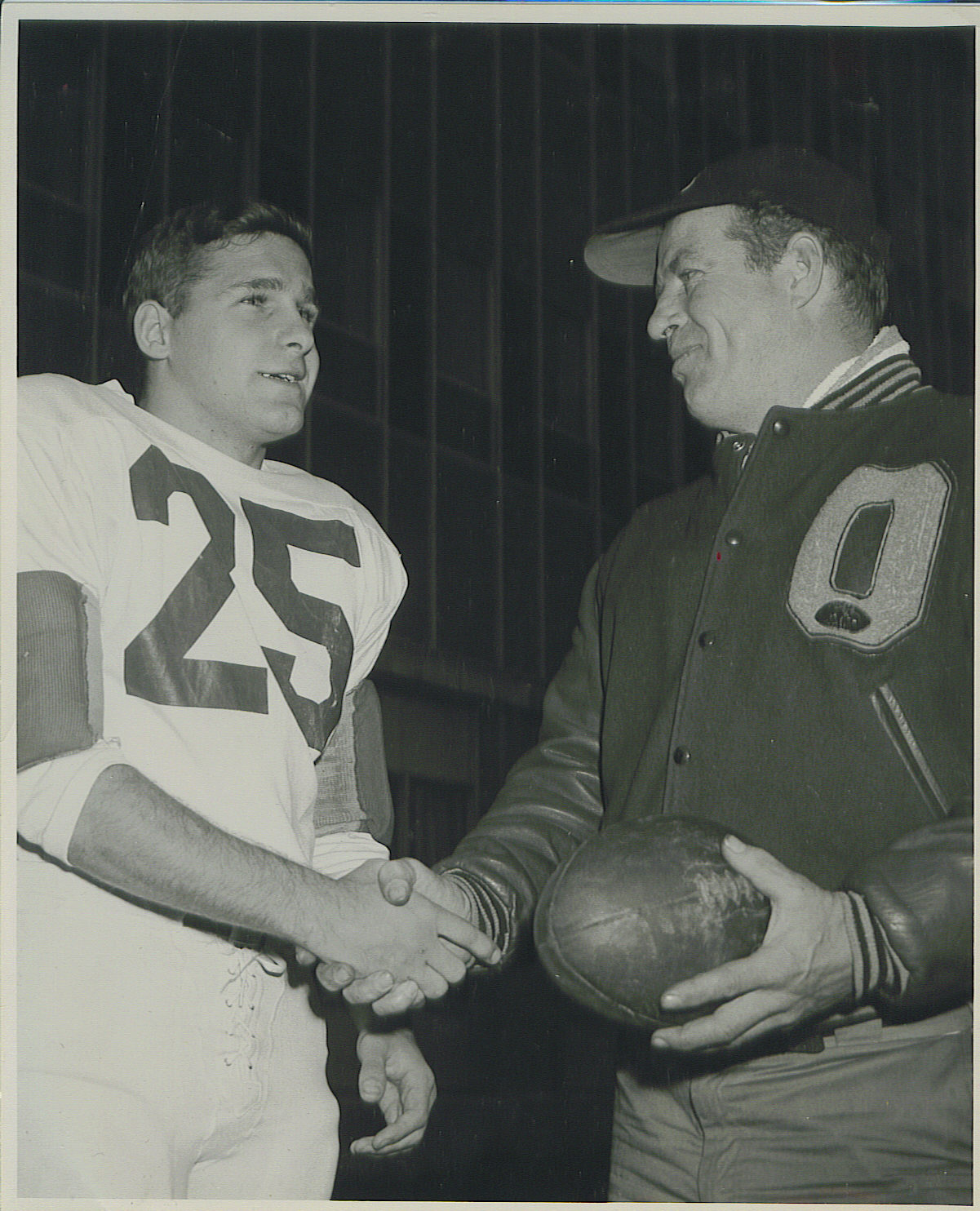 Player number 25 shakes hands with coach who is holding football.