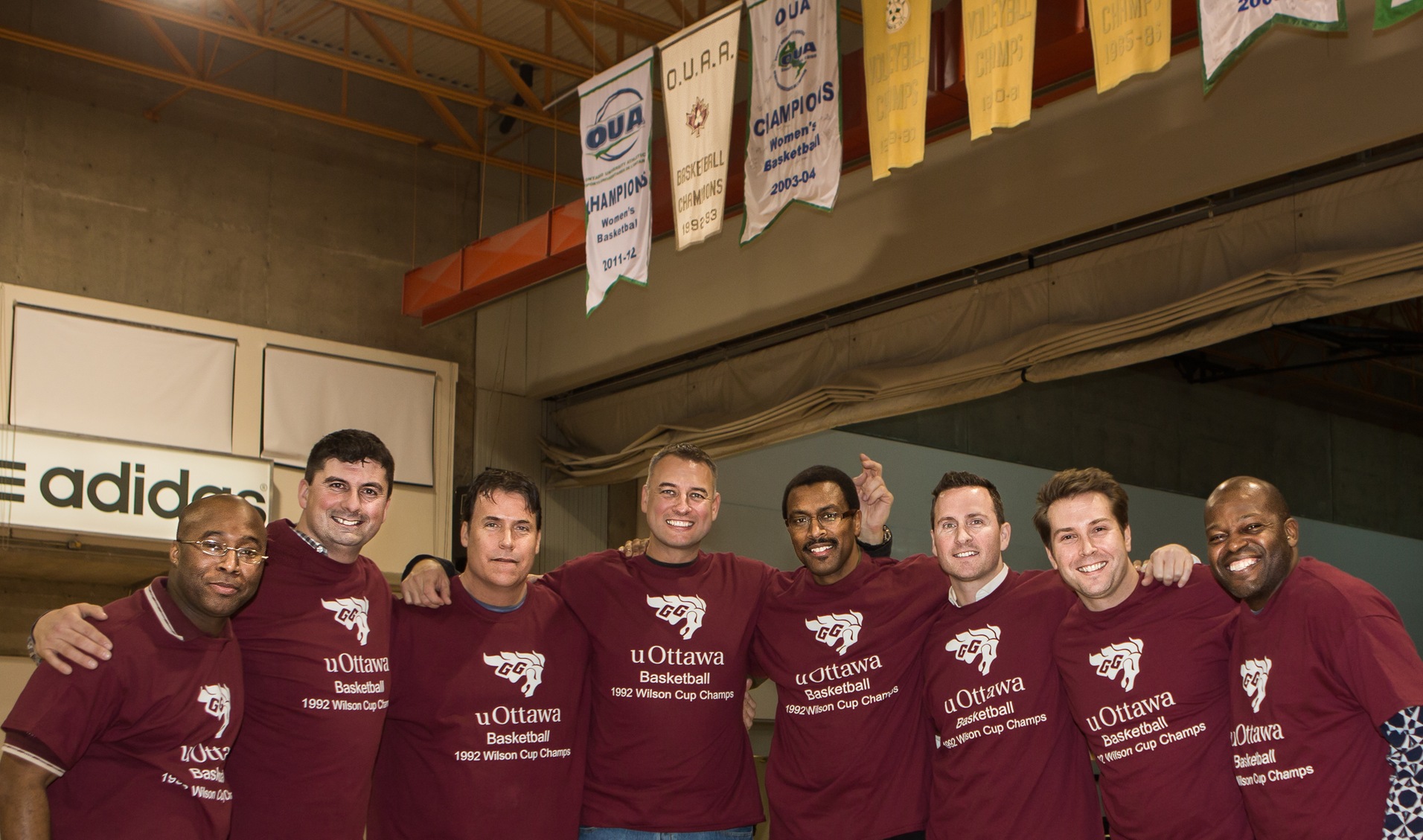 Group of nine alumni stand below the championship banner in the rafter of Montpetit Hall