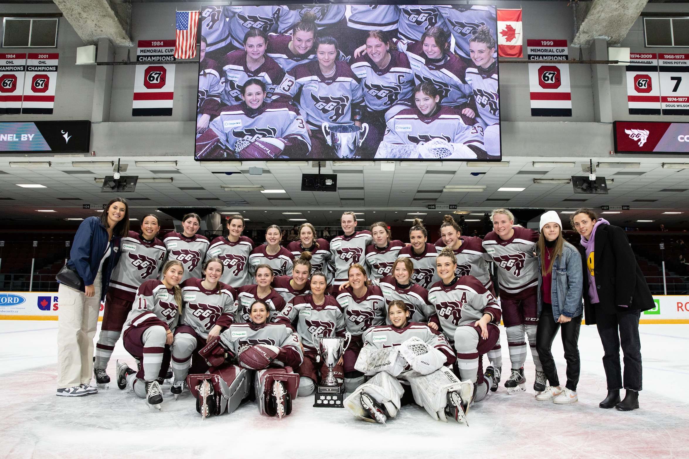 The Gee-Gees women's hockey team gathers around the Alerts Cup trophy on the ice at TD Place with the stadium TV screen behind them.