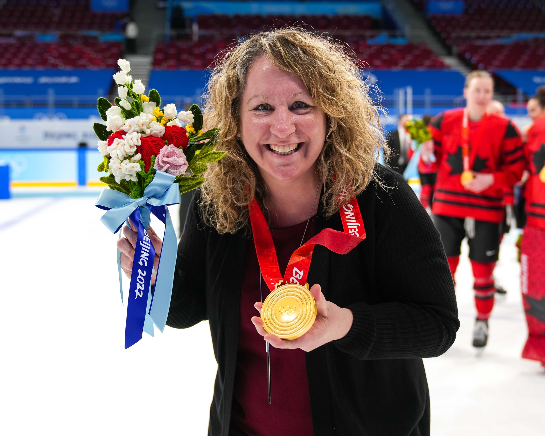 Kim Thompson holding her Olympic gold medal with players on ice behind her.
