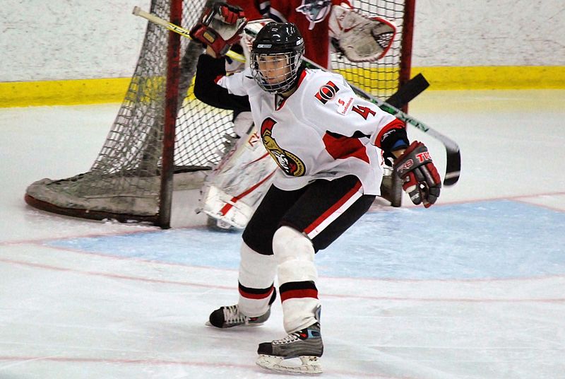 Mandi Duhamel playing ice hockey in a white Ottawa Senators, with the opposing team's goaltender in the background.