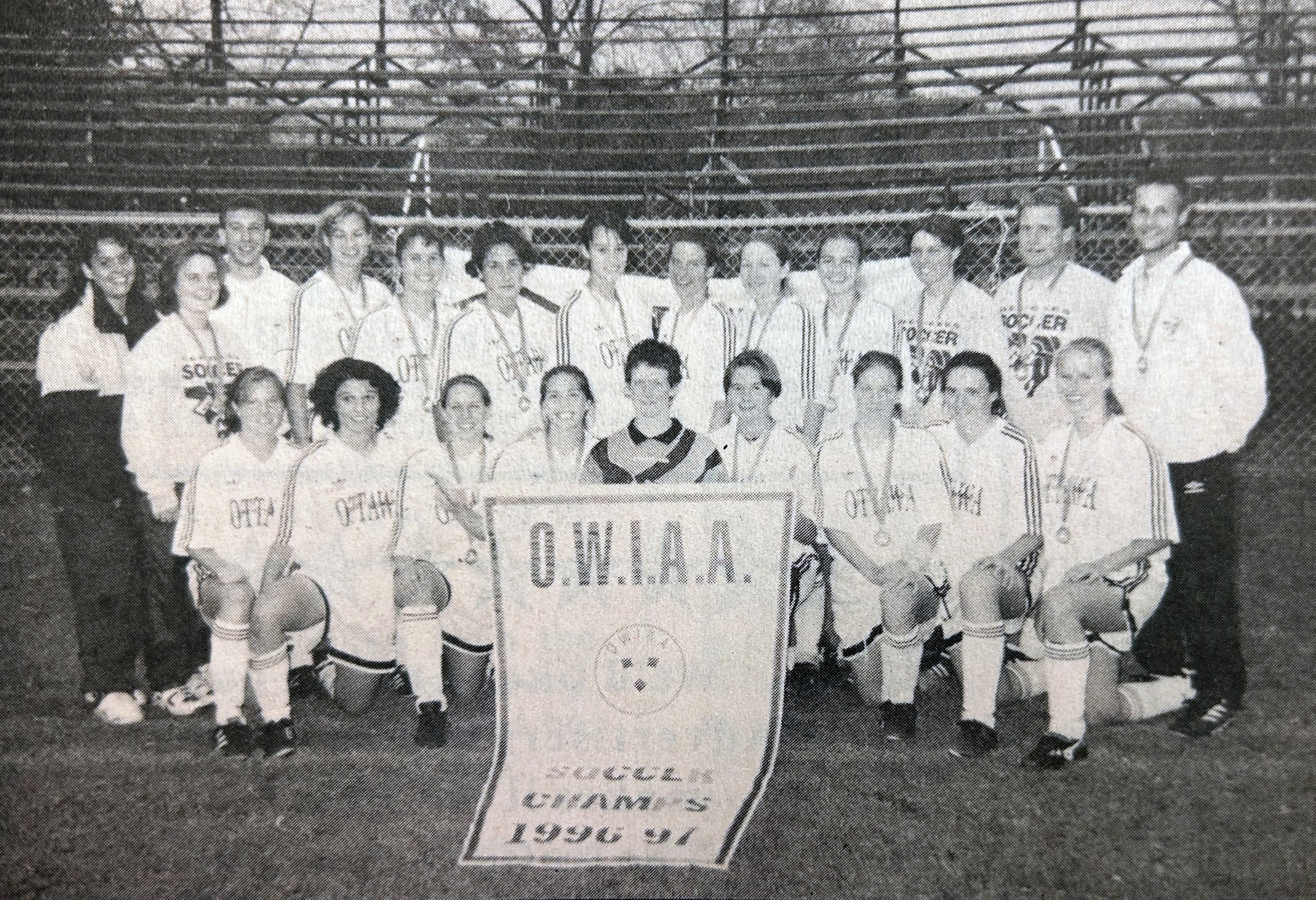 Black and white newspaper clipping of a the Gee-Gees women's soccer team with the OWIAA championship banner and their medals for 1996.